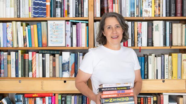 Eine Frau steht vor einem Bücherregal und hält einen Stapel Bücher im Arm.