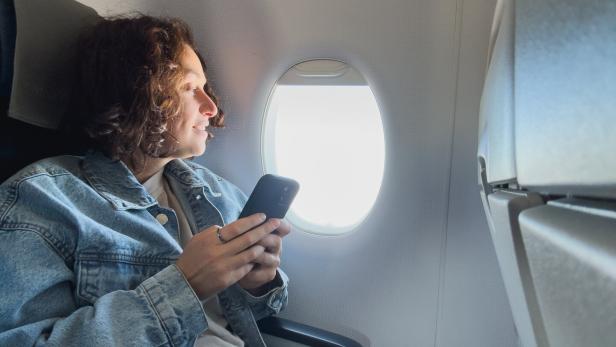Passenger woman is flying in plane. Girl using phone sitting by airplane window. Traveling female with smartphone inside plane enjoying flight. Woman using wifi connection on board. Traveling girl.