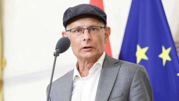 Ein Mann mit Schiebermütze und Brille spricht vor einer EU-Flagge in ein Mikrofon und hält einen Notizblock in der Hand.
