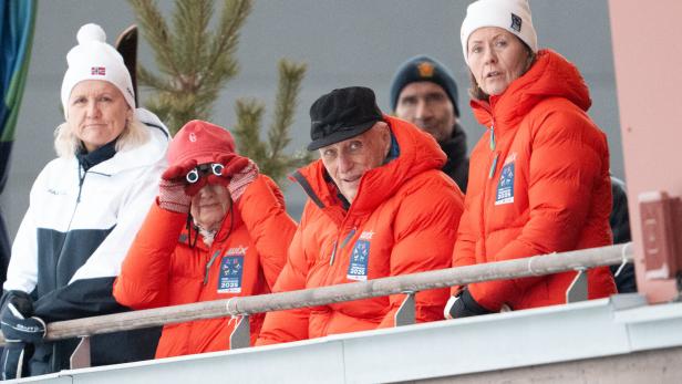 König Harald V. von Norwegen und Königin Sonja beobachten in orangefarbenen Jacken mit Trondheim 2025-Logo ein Sportereignis von einer Tribüne aus.