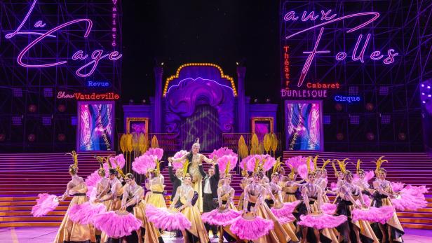 Bühnenensemble in goldenen Kostümen mit pinken Federn vor einer neonbeleuchteten Varieté-Showkulisse.