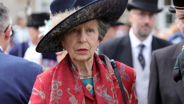 Prinzessin Anne trägt einen auffälligen roten Mantel mit Blumenmuster und einen dunklen Hut mit Federschmuck bei einer öffentlichen Veranstaltung.