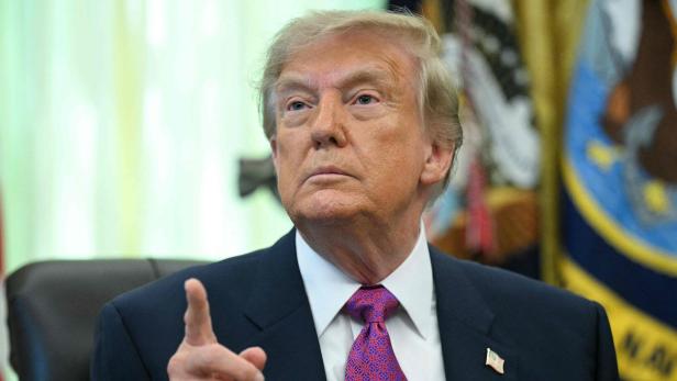 Donald Trump sitzt in einem Anzug mit Krawatte vor einer US-Flagge und hebt mahnend den Zeigefinger.