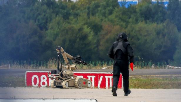 Ein Entschärfer in Schutzkleidung geht neben einem ferngesteuerten Roboter auf einem Flugfeld an einem roten Hinweisschild vorbei.