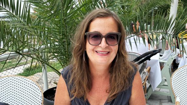 Ulli Sima sitzt, mit Sonnenbrille und breitem Grinsen, vor einer Palme und hält einen Teller mit Cesars Salad in den Händen