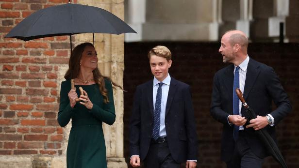 Prinzessin Kate, Prinz George und Prinz William gehen bei Regen gemeinsam vor einem Backsteingebäude spazieren.