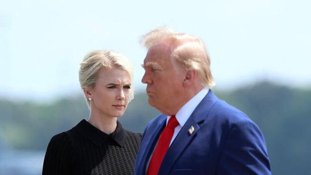Donald Trump in einem blauen Anzug geht neben einer blonden Frau in einem schwarzen Kleid im Freien.
