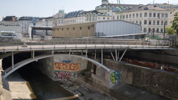 Eine Fußgängerbrücke überspannt den Wienfluss in Wien, umgeben von städtischer Architektur und mit Graffiti verzierten Betonmauern.