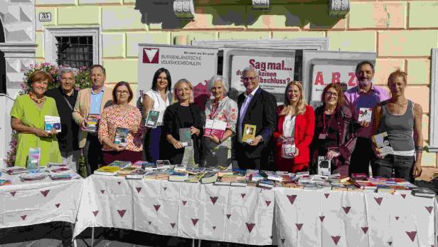Gruppe von zehn Personen steht vor Büchertischen und hält Bücher vor Werbebannern im Freien.