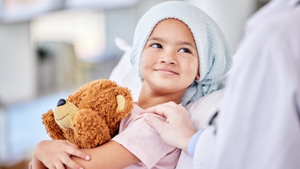 Ein lächelndes Kind mit Kopfbedeckung hält einen Teddybär und wird von einer Person sanft gestützt.