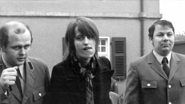 Ein schwarz-weiß Foto von Astrid Proll flankiert von zwei Männern am Rande des Prozesses im Jahr 1973
