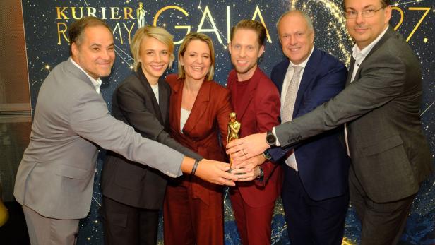 Sechs Personen halten gemeinsam eine goldene Romy-Trophäe vor einer „Kurier Romy Gala“-Wand.