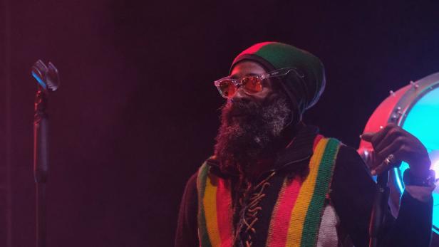 Marlon B. mit Rastafari-Mütze und buntem Pullover auf einer Bühne vor einem Mikrofon.