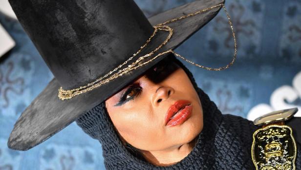 Porträt einer Person mit großem schwarzen Hut, Strickhaube und auffälligem Make-up, Blick schräg nach oben.
