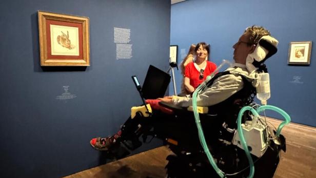 Person im Elektrorollstuhl mit Atemgerät betrachtet Kunst in einem Museum.