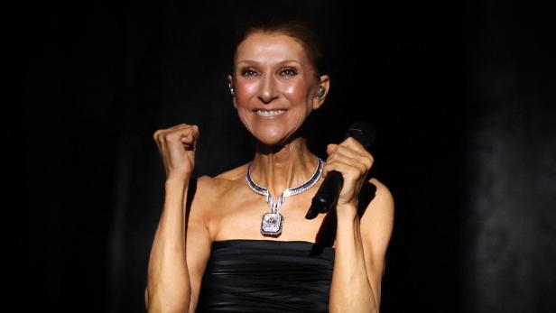 Céline Dion lächelt vor schwarzem Hintergrund, hält ein Mikrofon in der Hand und ballt die andere Faust vor der Brust.