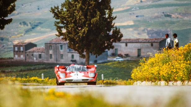 Ein roter Ferrari 312P mit der Startnummer 4 fährt auf einer ländlichen Straße an Zuschauern und gelben Blumen vorbei.