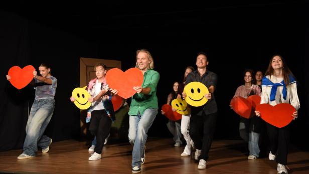 Eine Gruppe von Menschen steht auf einer Bühne vor schwarzem Hintergrund und hält rote Herzen sowie gelbe Smileys in die Kamera.