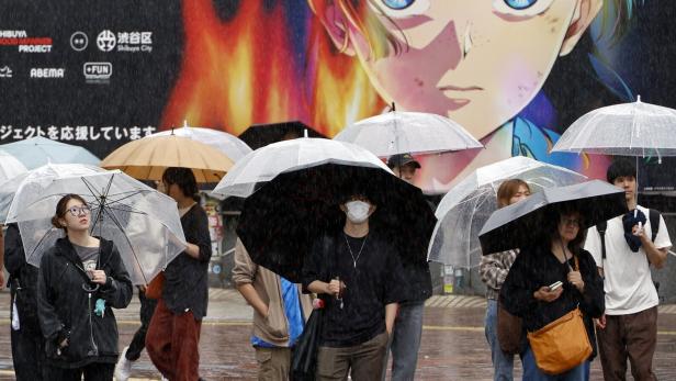 Menschen mit Regenschirmen gehen bei Regen an einer großen Werbetafel mit einem Anime-Gesicht in Shibuya City vorbei.