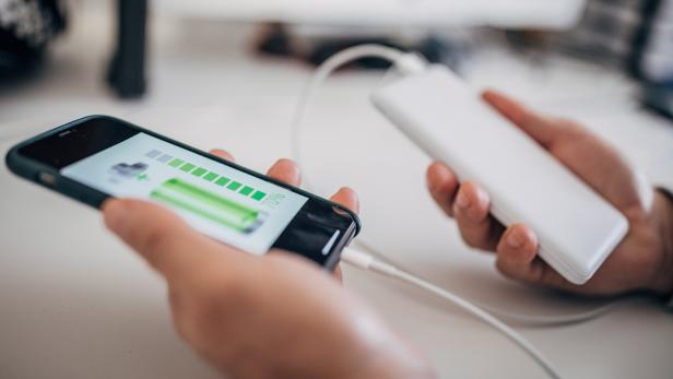 Eine Person hält ein Smartphone, das über ein Kabel mit einer Powerbank verbunden ist und einen Ladestand von 70 Prozent anzeigt.
