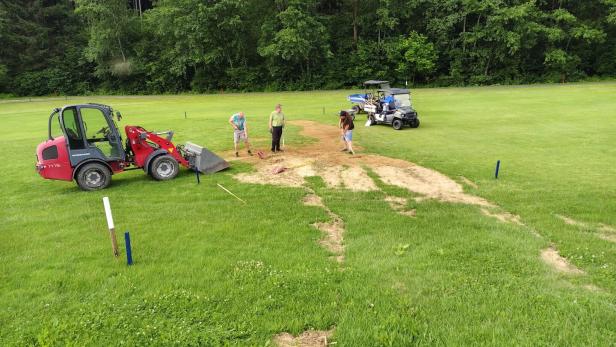 Drei Personen arbeiten auf einer Wiese mit Bagger und Golfcart an einer freigelegten Bodenstelle.