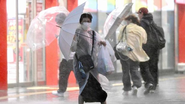 Person mit Maske hält im Regen einen transparenten Regenschirm und Einkaufstüten auf einer nassen Straße.
