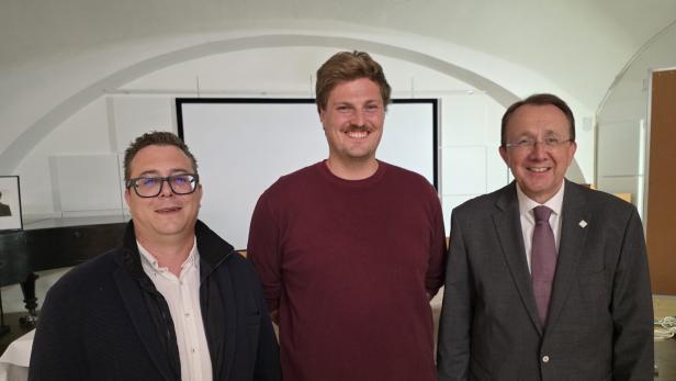 Drei lächelnde Männer stehen in einem Raum mit Projektionswand.