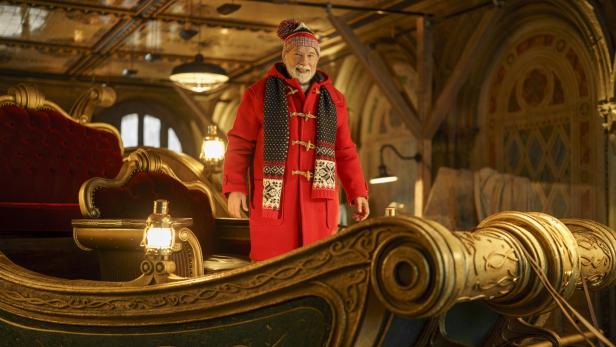 Arnold Schwarzenegger steht als Santa Clause in einem roten Dufflecoat und einer Wintermütze in einem prunkvollen, goldenen Schlitten.