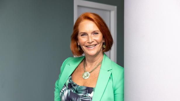 Lächelnde Frau mit rotem Bob, grünem Blazer und gemustertem Top lehnt an einer Wand.