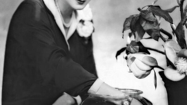 Schwarzweiß-Porträt einer elegant gekleideten Frau neben einem Goldfischglas und Blumen. Sie testet die Wasserdichtheit ihrer Rolex.