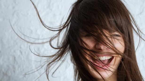 Lachende Frau mit vom Wind verwehten Haaren vor hellem Hintergrund.