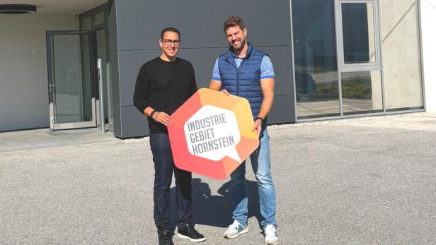 Zwei Männer stehen vor einem modernen Gebäude und halten ein Schild mit „Industriegebiet Hornstein“.