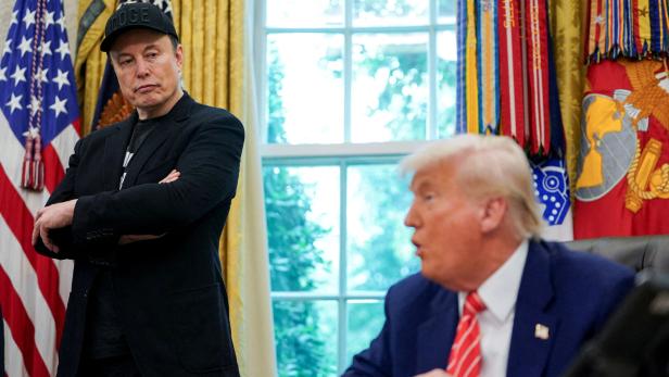 Elon Musk steht mit verschränkten Armen neben Donald Trump im Oval Office.