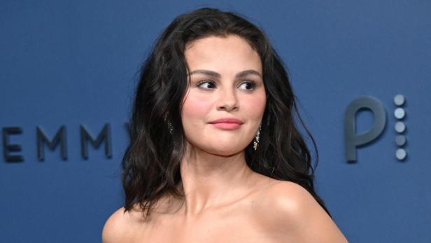 Selena Gomez posiert in einem trägerlosen, goldenen Kleid vor einem blauen Hintergrund mit EMMY-Schriftzug.