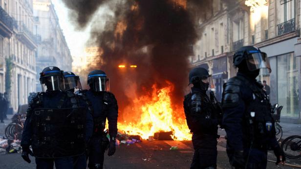 Polizisten in Schutzkleidung stehen vor einem brennenden Barrikadenfeuer in einer verqualmten Straße.
