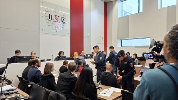 Mehrere Personen, Richter, Anwälte, Justizwachebeamte und Angeklagte sitzen und stehen in einem Gerichtssaal