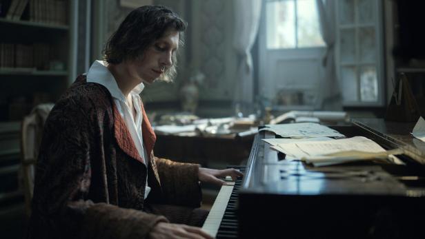 Ein langhaariger Mann sitzt in einem historischen Raum und spielt konzentriert Klavier.
