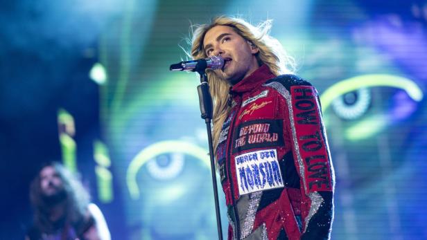 Bill Kaulitz singt mit blonden Haaren und einem bunten Bühnenoutfit in ein Mikrofon.