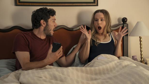 Adam Brody und Kristen Bell sitzen im Bett und reagieren überrascht auf etwas am Smartphone.