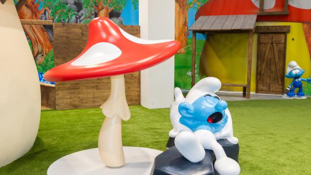 Schlumpf-Figuren in einer bunten Spielwelt mit großem roten Pilz und grasigem Boden.