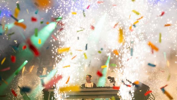 Ein DJ steht vor einem Pult, während im Hintergrund ein Feuerwerk und bunte Konfettistücke durch die Luft fliegen.