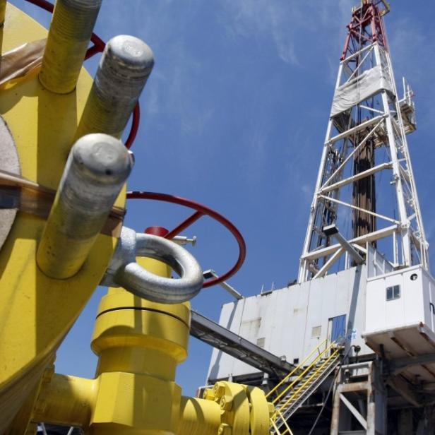 Ein Bohrturm ragt vor blauem Himmel in die Höhe, im Vordergrund gelbe Rohrleitungen.