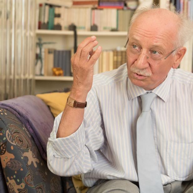 Ein älterer Mann mit Brille gestikuliert auf einem Sofa vor einem Bücherregal.