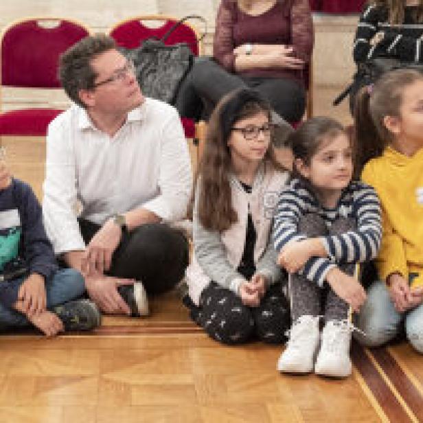 Eine Gruppe von Kindern sitzt mit einem Mann auf dem Boden und schaut aufmerksam nach oben.