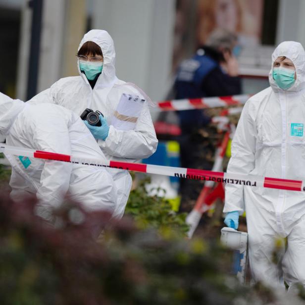 Drei Personen in Schutzanzügen, Masken und Handschuhen arbeiten an einem Tatort, der mit rot-weißem Band abgesperrt ist.