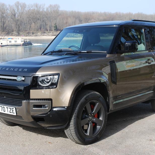 Ein brauner Land Rover Defender steht vor einem Fluss mit einer Fähre.