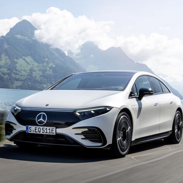 Ein weißer Mercedes-Benz EQS fährt auf einer Straße entlang eines Sees mit Bergen im Hintergrund.