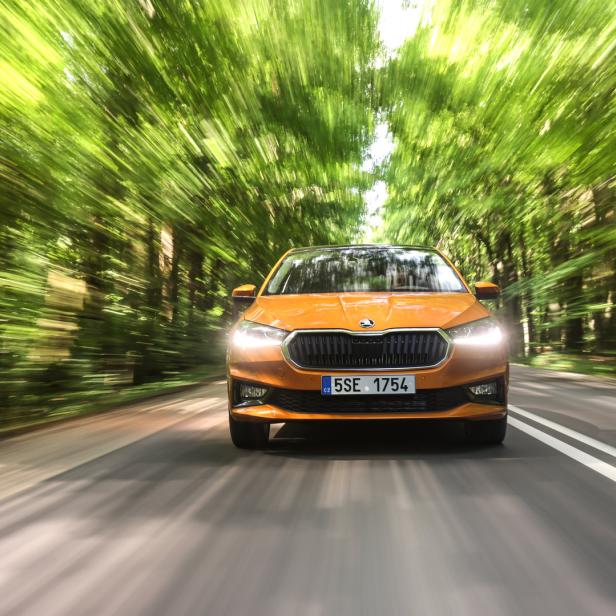 Ein orangefarbener Škoda fährt auf einer Straße durch einen grünen Wald.