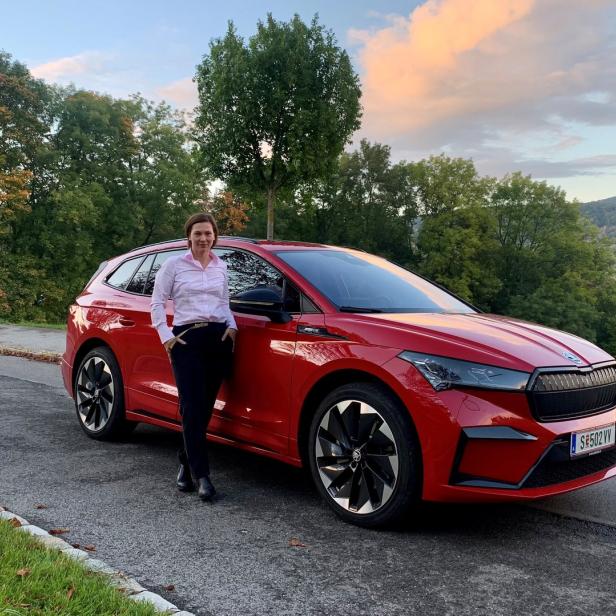 Eine Frau posiert neben einem roten Škoda Enyaq iV vor einer malerischen Landschaft.