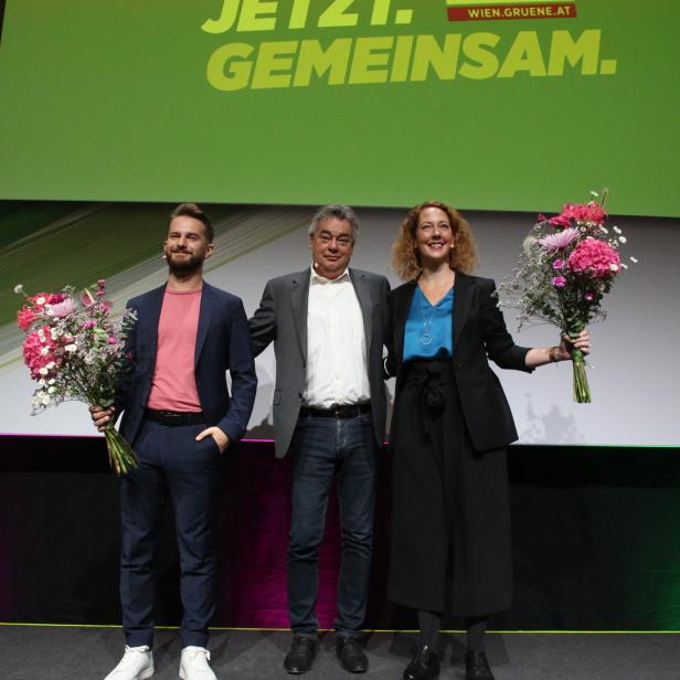 Drei Personen stehen mit Blumensträußen auf einer Bühne vor einem grünen Banner.